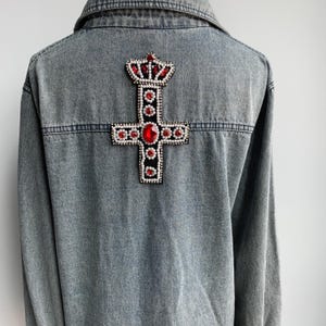 Może przedstawiać: Dżinsowa kurtka z dużym, zdobionym krzyżem i koroną na plecach. Krzyż wykonany jest z czarnej tkaniny z czerwonymi i białymi klejnotami.