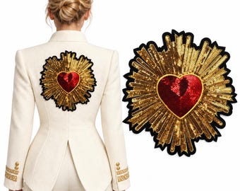 Op de afbeelding: Een crèmekleurige blazer met een grote, geborduurde patch op de rug. De patch toont een centraal rood hart van lovertjes omringd door stralende gouden stralen van lovertjes, omlijnd met een dikke zwarte rand op de stof.