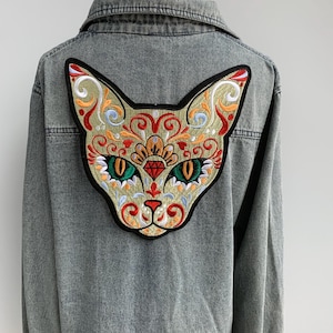 May include: A grey denim jacket with an embroidered cat face on the back. The cat design is detailed with red, orange, green, and gold thread, outlined in black. The jacket has a classic collar and a button-down front.