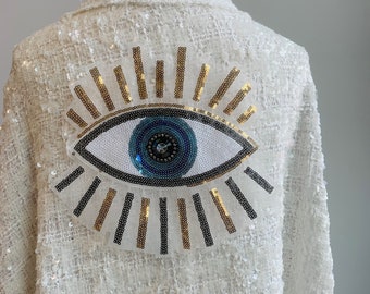 May include: White sequined jacket with a large blue and white evil eye design on the back. The eye is surrounded by gold and black sequins.