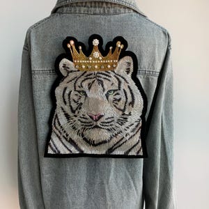 May include: A denim jacket with a white tiger embroidered patch on the back. The tiger is wearing a gold crown with white jewels.