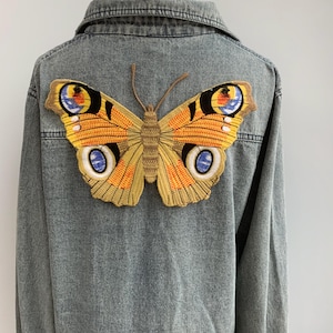 以下が含まれることがあります： 背中に大きな蝶の刺繍パッチが付いた、薄いブルーのデニムジャケット。 蝶はオレンジ、黄色、青、茶色を使用しています。