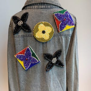 Könnte beinhalten: Eine graue Jeansjacke mit fünf gestickten Patches. Die Patches haben die Form einer Blume und sind mit bunten Perlen verziert. Die Patches sind schwarz, gelb, rot, grün und lila.