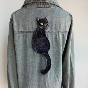 Può includere: Toppa ricamata nera da stirare con un gatto nero con occhi verdi. Il gatto è seduto con la coda arrotolata attorno al suo corpo.