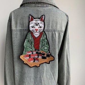 以下が含まれることがあります： 白い猫が緑と赤の着物と白いヘッドバンドを着てデニムジャケットに刺繍されています。猫は寿司の皿を持っています。