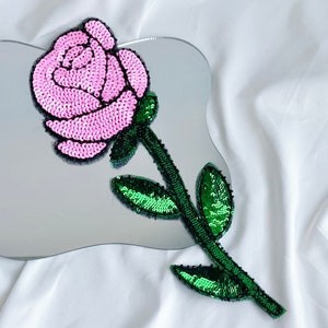 Könnte beinhalten: Eine rosa und grüne Pailletten-Rosenapplikation. Die Rose besteht aus rosa Pailletten und der Stiel und die Blätter aus grünen Pailletten.