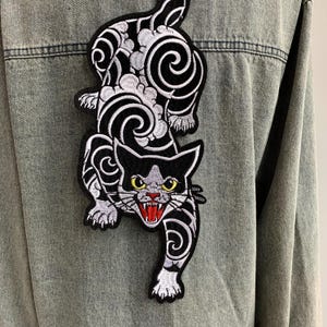 Peut inclure: Une veste en jean gris clair avec un grand &eacute;cusson brod&eacute; au dos. L'&eacute;cusson repr&eacute;sente un chat noir et blanc aux yeux jaunes, la gueule ouverte montrant des gencives rouges et des dents blanches, entour&eacute; de motifs de nuages tourbillonnants.