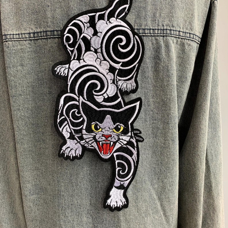 Peut inclure: Une veste en jean gris clair avec un grand &eacute;cusson brod&eacute; au dos. L'&eacute;cusson repr&eacute;sente un chat noir et blanc aux yeux jaunes, la gueule ouverte montrant des gencives rouges et des dents blanches, entour&eacute; de motifs de nuages tourbillonnants.