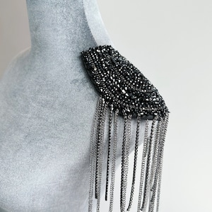 Pode incluir: Peça de ombro preta com contas e franjas de correntes prateadas.