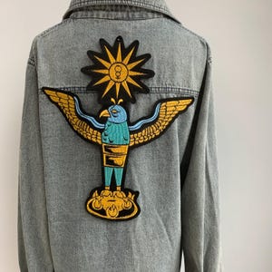 May include: A denim jacket with an embroidered patch of a blue bird with outstretched wings, a yellow sun with a symbol in the center, and a yellow flame design at the base.