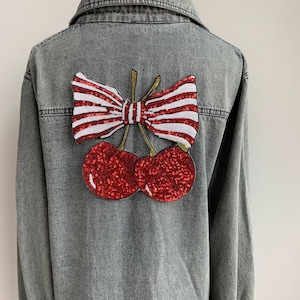 May include: Gray denim jacket with a sequined cherry and bow design on the back. The cherries are red, and the bow is red and white striped. The jacket has a classic collar and button-up front.