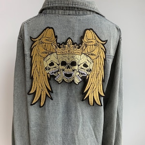 Pode incluir: Jaqueta jeans cinza com um grande patch dourado bordado nas costas. O patch mostra três caveiras usando uma coroa, com asas douradas. A jaqueta tem fechamento por botões e gola.