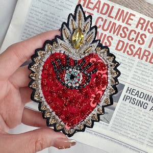Può includere: Un cerotto ricamato a forma di cuore rosso con un grande occhio al centro. L'occhio è circondato da paillettes e perline. Il cuore è bordato con filo dorato e ha una corona dorata in cima.