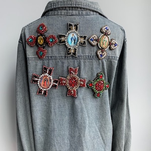 Puede incluir: Una chaqueta vaquera con una variedad de adornos coloridos en forma de cruz. Las cruces están decoradas con pedrería, cuentas e imágenes de figuras religiosas.