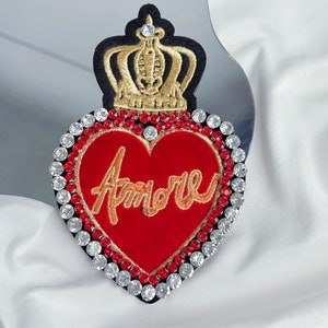 Pode incluir: Um coração de feltro vermelho com um "Amore" bordado em ouro rodeado por strass transparentes e vermelhos. Uma coroa dourada com strass fica em cima do coração.