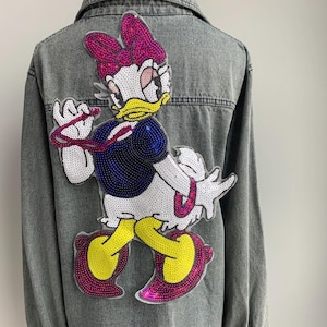 Könnte beinhalten: Eine graue Jeansjacke mit einem gestickten Pailletten-Aufnäher eines Comic-Entchens mit einer rosa Schleife, gelben Schuhen und einem blauen Kleid.