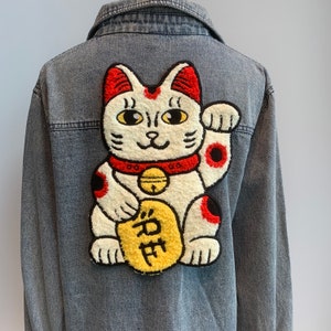 Peut inclure: Une veste en jean avec un grand patch brodé représentant un Maneki-Neko, un chat porte-bonheur japonais, blanc, rouge et noir, tenant une pièce d'or avec le caractère japonais pour "fortune" dessus.