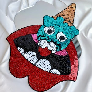 May include: A red, black, and white sequined patch of a cartoon monster wearing a cone-shaped hat. The monster has large, round eyes and a wide, open mouth.