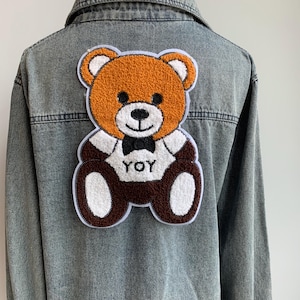 May include: A denim jacket with a large, embroidered teddy bear patch on the back. The bear is brown with a white belly and a black bow tie. The text "YOY" is on the bear's shirt.