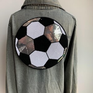 May include: A denim jacket with a large, round soccer ball design on the back. The soccer ball is black and white with silver sequins. The jacket is a light gray color.