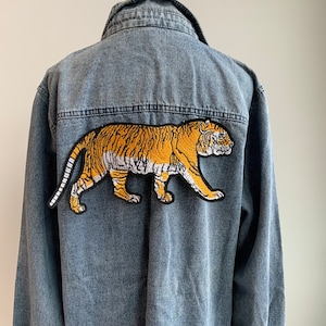 May include: A faded blue denim jacket with a large embroidered tiger patch on the back. The tiger design is in orange, black, and white, with detailed stitching. The jacket has a classic collar and a button-down front.