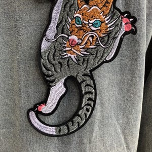 Peut inclure: Une veste en jean gris orn&eacute;e d'un grand &eacute;cusson brod&eacute; repr&eacute;sentant un tigre stylis&eacute; avec une fourrure orange, des yeux bleu sarcelle et des touches roses sur les pattes et le museau. Le tigre est repr&eacute;sent&eacute; dans une posture courb&eacute;e.
