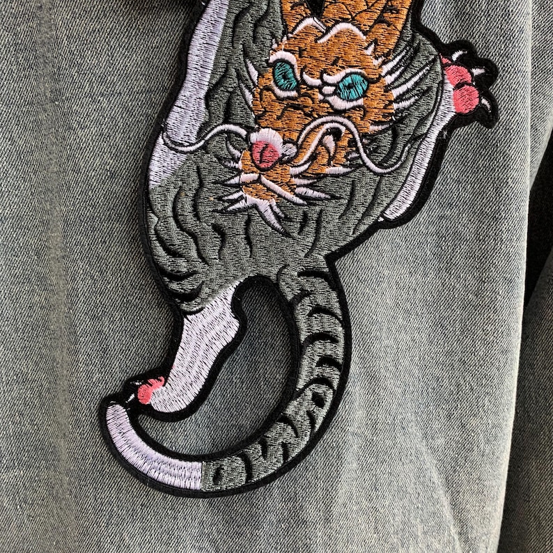 Peut inclure: Une veste en jean gris orn&eacute;e d'un grand &eacute;cusson brod&eacute; repr&eacute;sentant un tigre stylis&eacute; avec une fourrure orange, des yeux bleu sarcelle et des touches roses sur les pattes et le museau. Le tigre est repr&eacute;sent&eacute; dans une posture courb&eacute;e.