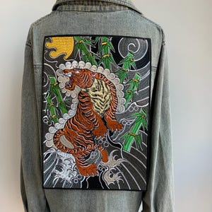 Könnte beinhalten: Graue Jeansjacke mit einem großen, aufgestickten Aufnäher auf dem Rücken. Der Aufnäher zeigt einen brüllenden Tiger in Braun-, Orange- und Gelbtönen, vor dem Hintergrund von Bambus und wirbelndem Wasser.
