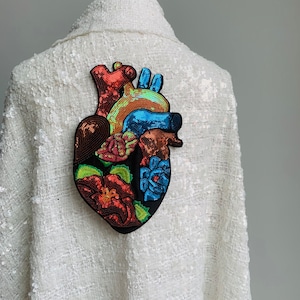 May include: A white sequined jacket with a colourful embroidered heart patch. The heart is made of sequins and features a floral design.