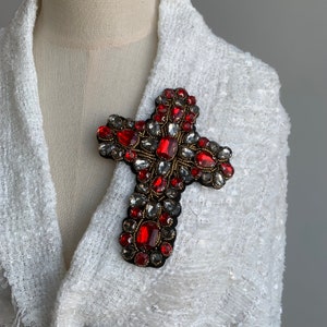 May include: A large, red and black jeweled cross brooch. The cross is made of rhinestones and beads, and it is attached to a white fabric background.