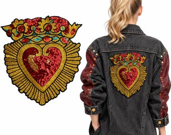 Pode incluir: Um casaco de ganga preta com mangas vermelho-escuras com tachas metálicas e um grande emblema bordado em forma de coração nas costas. O emblema apresenta um coração de lantejoulas vermelhas com uma coroa floral e raios dourados.