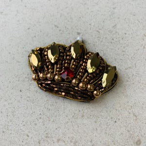 Op de afbeelding: Een goud- en bruin kralen kroonbroche met een rood juweel in het midden. De kroon is versierd met goudkleurige druppelvormige edelstenen en kleine gouden kralen. Bovenop zit een kleine witte parel.