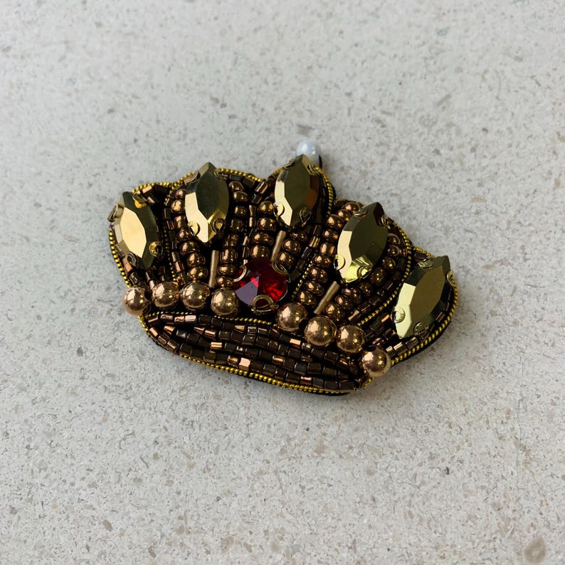 Op de afbeelding: Een goud- en bruin kralen kroonbroche met een rood juweel in het midden. De kroon is versierd met goudkleurige druppelvormige edelstenen en kleine gouden kralen. Bovenop zit een kleine witte parel.