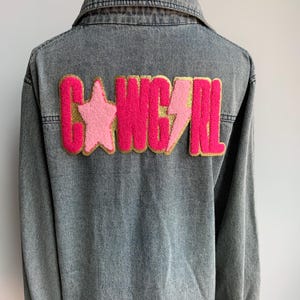 May include: A denim jacket with a pink and gold "COWGIRL" patch on the back. The patch features a star, lightning bolt, and glitter accents.