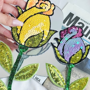 May include: Two embroidered iron-on patches featuring colorful sequined roses. The roses are yellow and purple with green leaves. The patches are perfect for adding a touch of whimsy to clothing, bags, or other fabric items.