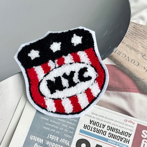 May include: A red, white and blue felt patch with a white circle in the centre that says "NYC" in black letters. The patch is shaped like a shield with stars in the top corners.