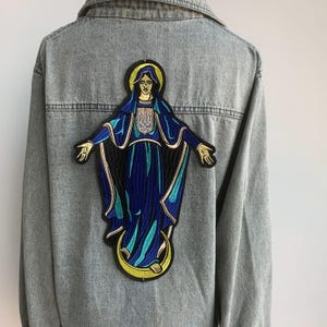 May include: A light-wash denim jacket featuring a large embroidered patch on the back. The patch shows a religious figure in blue robes with a gold halo and a Ukrainian coat of arms. The jacket has a classic collar and button-down front.