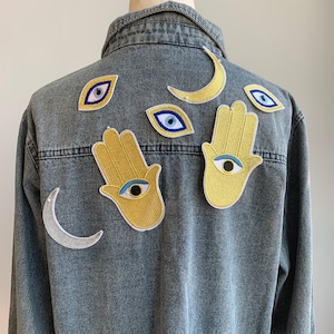 May include: A denim jacket with embroidered patches of a crescent moon, hamsa hands, and evil eyes. The patches are in shades of blue, yellow, and white.