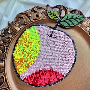 Puede incluir: Una aplicación de lentejuelas de color melocotón con un tallo y hojas bordadas en verde. La aplicación está hecha de lentejuelas en tonos de rosa, amarillo y rojo.
