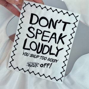 May include: A white fabric patch with black embroidered text that reads "DON'T SPEAK LOUDLY" and "YOU SHUP TOO NOISY OFF!". The patch has a black zigzag border.