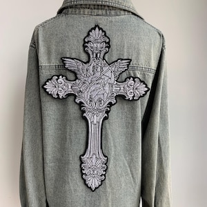 May include: A grey denim jacket with a large, ornate cross patch on the back. The patch features a detailed design of an angel with wings, rendered in shades of grey and black. The jacket has a classic button-up design.