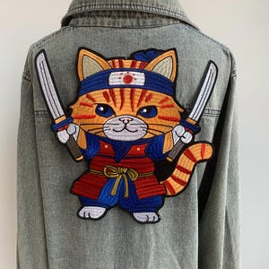 以下が含まれることがあります： 二本の刀を持った侍に扮した漫画の猫の刺繍パッチ。猫はオレンジと白の毛皮、青い目を持ち、赤い丸の付いた青いヘアバンドを着用しています。パッチはデニムジャケットに取り付けられています。