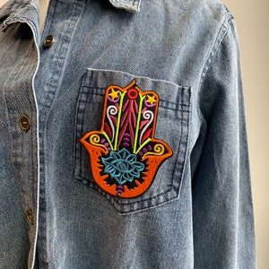 Könnte beinhalten: Ein Jeanshemd mit einem bunten gestickten Hamsa-Hand-Patch auf der Tasche. Die Hamsa-Hand ist ein Symbol für Schutz und Glück.