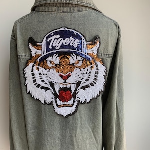 Könnte beinhalten: Graue Jeansjacke mit einem großen Pailletten-Tigerkopf-Aufnäher auf dem Rücken. Der Tiger-Aufnäher zeigt eine blaue Baseballkappe mit dem Wort "Tigers" in weißer Schrift. Das Gesicht des Tigers ist orange, weiß und rot.