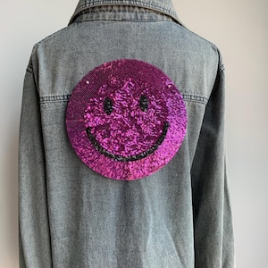 Puede incluir: Una chaqueta vaquera con un gran parche de smiley de lentejuelas rosas en la espalda.