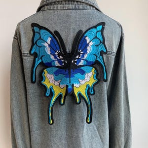 May include: A blue denim jacket with a large embroidered butterfly on the back. The butterfly is blue, yellow, and white with a dripping effect.