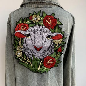 May include: A light-wash denim jacket featuring a large embroidered patch on the back. The patch depicts a sheep's face surrounded by red roses, green leaves, and yellow flowers. The jacket has a classic collar and button-up front.