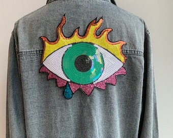 Könnte beinhalten: Eine Jeansjacke mit einem großen, bunten Augen-Applikationen auf der Rückseite. Das Auge ist von Flammen umgeben und hat eine Träne darunter.