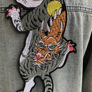 Peut inclure: Un grand &eacute;cusson brod&eacute; repr&eacute;sentant un tigre gris et un dragon orange entrelac&eacute;s, cousu sur une veste en jean bleu clair. Le tigre a des griffes roses et le dragon a des yeux bleu sarcelle, un nez rose et un soleil jaune derri&egrave;re lui.