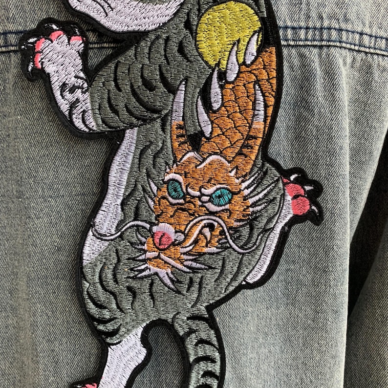 Peut inclure: Un grand &eacute;cusson brod&eacute; repr&eacute;sentant un tigre gris et un dragon orange entrelac&eacute;s, cousu sur une veste en jean bleu clair. Le tigre a des griffes roses et le dragon a des yeux bleu sarcelle, un nez rose et un soleil jaune derri&egrave;re lui.
